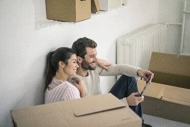 Ehepaar sitzt in seinem neuen Haus, umgeben von Kartons, und schaut auf ein Tablet - MOEF00683
