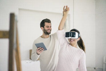 Man lifting woman's arm wearing VR glasses in new home - MOEF00689