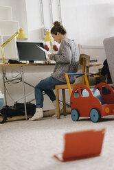 Frau sitzt am Schreibtisch zu Hause umgeben von Spielzeug - KNSF03418