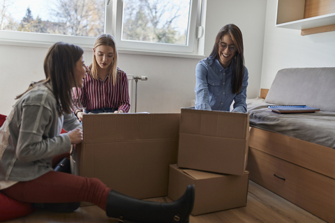 Drei glückliche junge Frauen beim Auspacken von Kartons in einem Zimmer, lizenzfreies Stockfoto