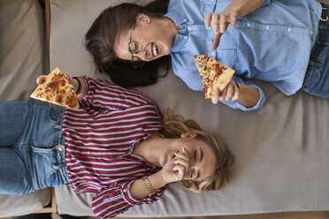 Two laughing young women lying down eating pizza together - ZEDF01096