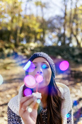 Woman wearing wooly hat blowing soap bubbles in an autumnal forest - MGOF03706