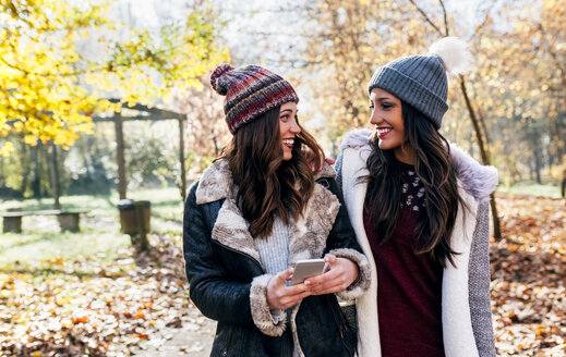 Zwei hübsche Frauen mit Handy lächeln sich in einem herbstlichen Wald an - MGOF03726