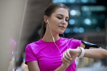 Junge Frau im rosa Sporthemd hört Musik und schaut auf ihre Smartwatch - SBOF00979