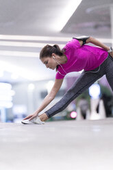 Young woman in pink sportshirt listening to music and stretching in front of modern building - SBOF00990