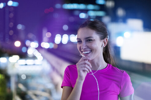 Smiling young woman in pink sportshirt listening to music in city at night - SBOF00994