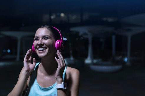 Glückliche junge Frau mit rosafarbenen Kopfhörern, die nachts in einer modernen städtischen Umgebung Musik hört - SBOF01024