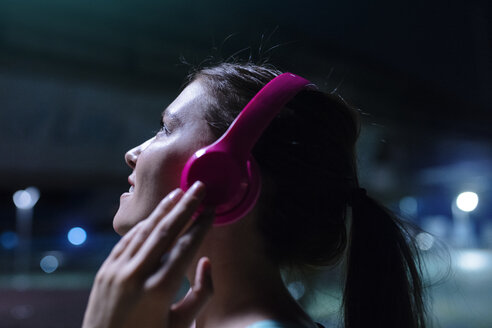 Glückliche junge Frau mit rosafarbenen Kopfhörern, die nachts in einer modernen städtischen Umgebung Musik hört - SBOF01025