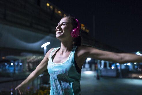 Glückliche junge Frau mit rosafarbenen Kopfhörern, die nachts in einer modernen städtischen Umgebung Musik hört - SBOF01026