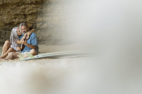 Handsome senior couple sitting on the beach, man playing guitar - SBOF01029