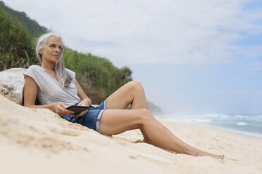 Schöne lächelnde ältere Frau am Strand liegend, hält E-Book - SBOF01089