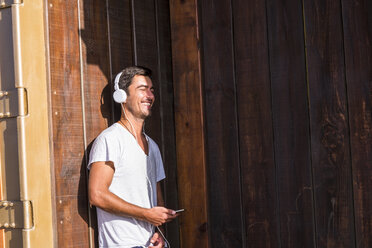 Happy young man with cell phone and headphones leaning against wooden wall - SIPF01915