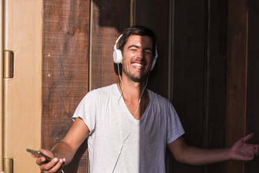 Happy young man with cell phone and headphones at wooden wall - SIPF01928