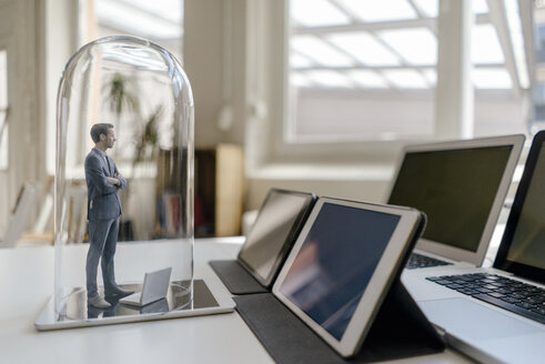 Businessman figurine standing under glass bell on desk, facing mobile devices - FLAF00040