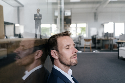 Geschäftsmann sitzt in seinem Büro mit seinem 3D-Porträt hinter einer Glasscheibe, lizenzfreies Stockfoto