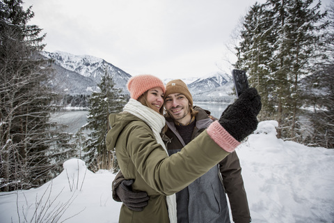 Lächelndes Paar macht ein Selfie in alpiner Winterlandschaft mit See, lizenzfreies Stockfoto