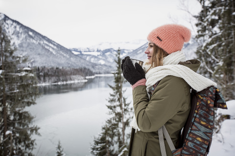 Young woman with hot drink standing in alpine winter landscape with lake stock photo