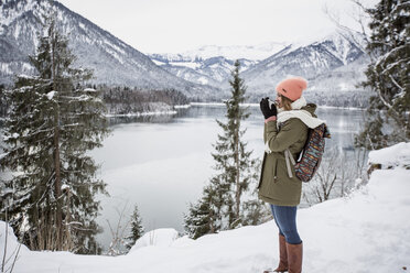 Young woman with hot drink standing in alpine winter landscape with lake - SUF00404