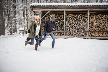 Glückliches Paar läuft im Winter vor einem Holzstapel im Freien - SUF00424