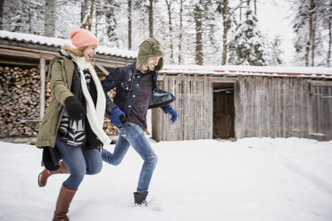 Happy couple running in front of wood pile outdoors in winter - SUF00425