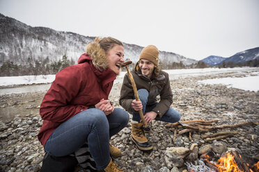 Happy couple on a trip in winter eating a sausage at camp fire - SUF00451