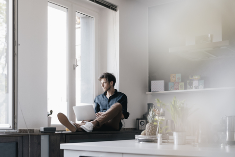 Young man with laptop sitting on window sill in a loft looking out of the window stock photo