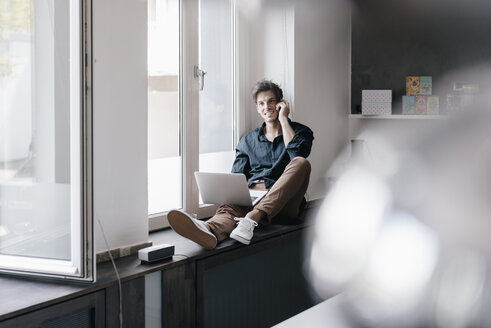 Smiling young man on the phone sitting with laptop on window sill in a loft - KNSF03421