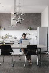 Junger Mann sitzt am Tisch in der Küche und arbeitet am Laptop - KNSF03437