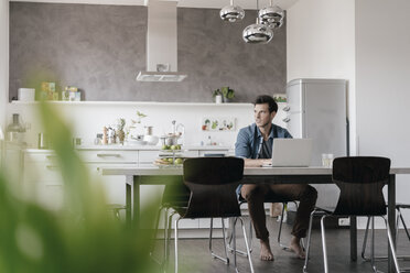 Young man sitting at table in the kitchen working on laptop - KNSF03438