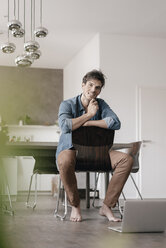 Portrait of smiling young man sitting on chair in a loft - KNSF03439