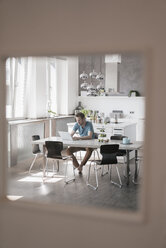 Man sitting at table in the kitchen working on laptop - KNSF03445