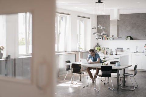 Man sitting at table in the kitchen working on laptop stock photo