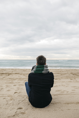 Rückenansicht eines Mannes, der im Winter am Strand sitzt und in die Ferne schaut, lizenzfreies Stockfoto