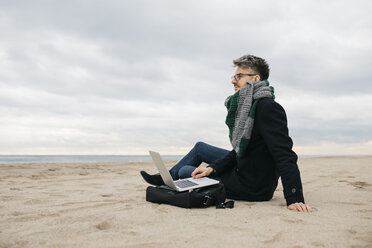 Businessman with laptop sitting on the beach in winter - JRFF01511