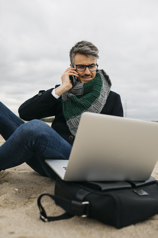 Porträt eines Geschäftsmannes am Telefon, der im Winter am Strand sitzt und einen Laptop benutzt, lizenzfreies Stockfoto