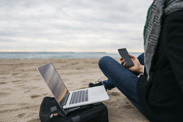 Businessman sitting on the beach in winter using laptop and cell phone, partial view - JRFF01513