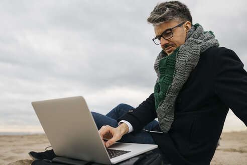 Geschäftsmann, der im Winter am Strand sitzt und einen Laptop benutzt - JRFF01514