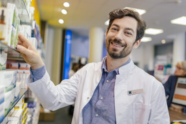 Portrait of smiling pharmacist with medicine at shelf in pharmacy - MFF04284