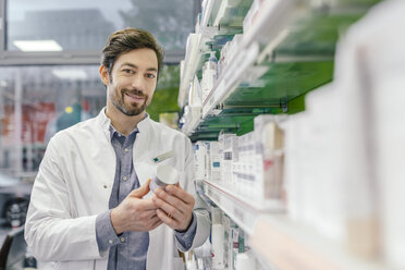 Porträt einer lächelnden Apothekerin mit Cremetiegel im Regal einer Apotheke - MFF04295