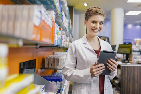 Porträt einer lächelnden Apothekerin am Regal mit Medikamenten in der Apotheke, die eine Tablet hält - MFF04347