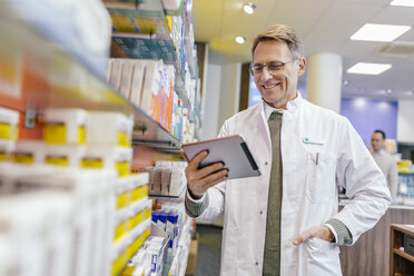 Smiling pharmacist at shelf with medicine in pharmacy holding tablet - MFF04350