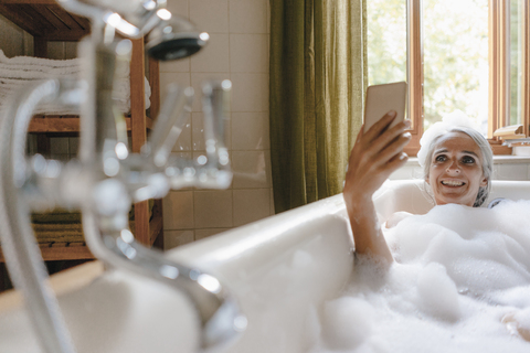 Porträt einer glücklichen Frau in der Badewanne, die ein Selfie mit ihrem Handy macht, lizenzfreies Stockfoto