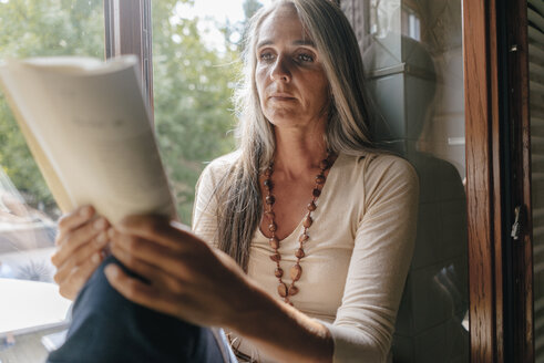 Porträt einer Frau, die zu Hause auf der Fensterbank sitzt und ein Buch liest - KNSF03470
