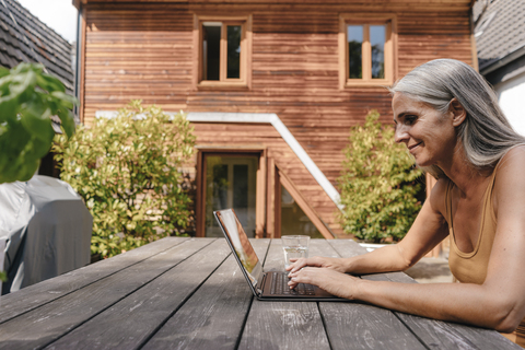 Frau sitzt auf der Terrasse ihres Hauses und benutzt einen Laptop, lizenzfreies Stockfoto