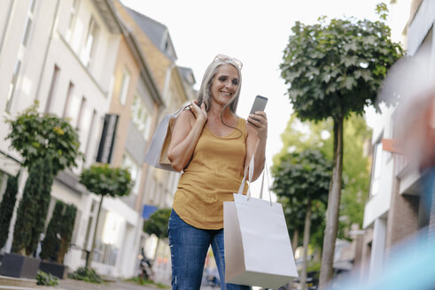 Lächelnde Frau auf der Straße, die Einkaufstüten trägt und auf ihr Handy schaut - KNSF03490