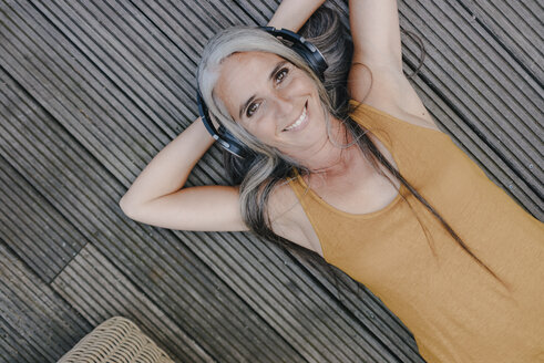 Porträt einer glücklichen Frau, die auf einer Terrasse liegt und mit Kopfhörern Musik hört - KNSF03493