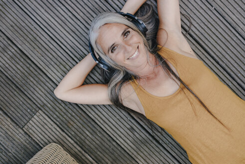 Portrait of happy woman lying on terrace listening music with headphones - KNSF03493