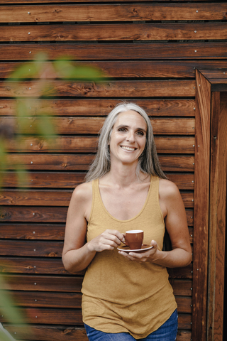 Porträt einer glücklichen reifen Frau mit Kaffeetasse vor einer Holzfassade, lizenzfreies Stockfoto
