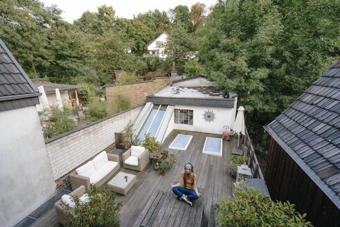 Frau mit Kopfhörern, die auf einer Terrasse sitzt und Yogaübungen macht - KNSF03536