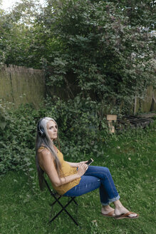 Frau mit Handy sitzt im Garten und hört Musik mit Kopfhörern - KNSF03539